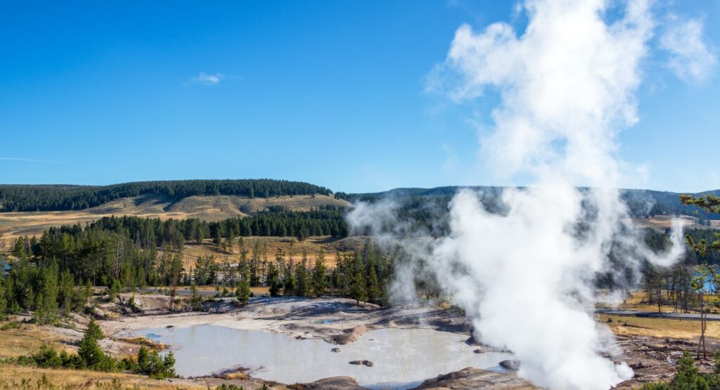 Expansive view of the Mud Volcano area in Yellowstone National Park with steam rising dramatically from a geothermal pool, surrounded by a rugged landscape of pine forests and rolling hills under a clear blue sky.