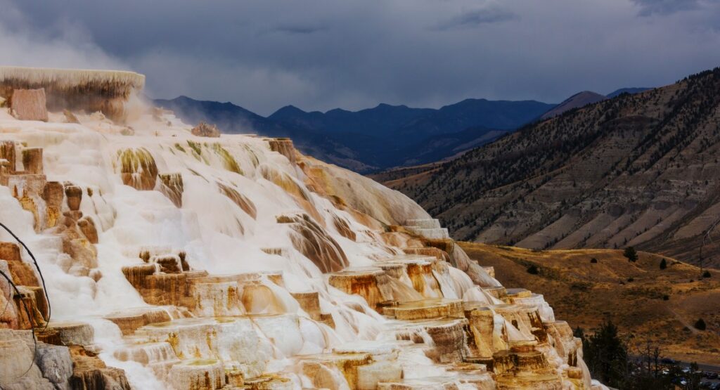 Majestic Mammoth Hot Springs terrace formations with steam rising against a dramatic mountain backdrop and stormy skies, highlighting the natural geothermal wonders of Yellowstone National Park