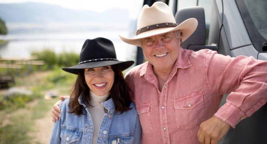 Cheerful couple in stylish cowboy hats smiling and posing for a photo beside their van with a scenic lake and greenery in the background