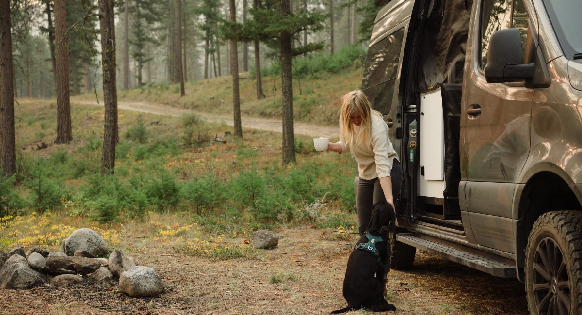  A woman enjoying a peaceful morning with her dog beside a camper van in a serene forest setting, with the warmth of a cup in her hands highlighting a tranquil escape into nature