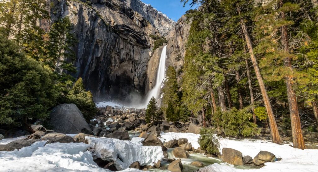 Lower Yosemite Falls in winter, captured in a long exposure that beautifully illustrates the waterfall's cascade amidst snow-covered rocks and evergreen trees