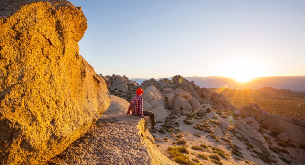 A hiker in a red beanie and plaid shirt sits on a rocky outcrop, taking in the breathtaking view of the sun setting over the expansive, rocky desert landscape in one of the national parks near Los Angeles