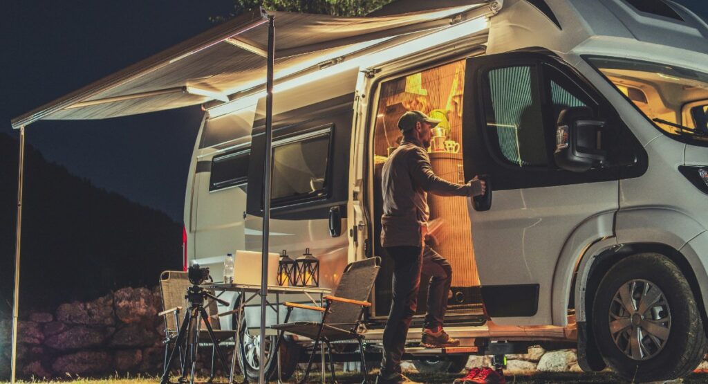 An adventurer sets up camp for the night with a modern camper van, awning extended for shelter, amidst the tranquil backdrop of a forest