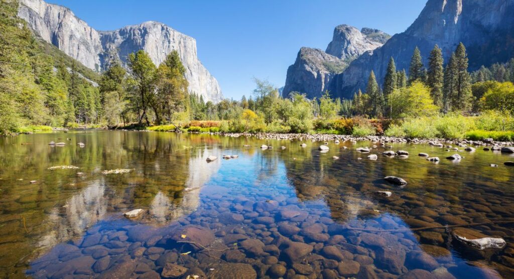 Crystal clear waters of the Merced River with a serene view of Yosemite Valley, El Capitan, and Half Dome in the background, reflecting the lush greenery and clear blue skies