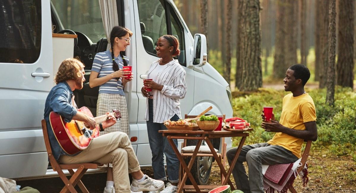 Multi-generational family enjoying outdoor dining setup with folding table and chairs beside Class B campervan in forest campground