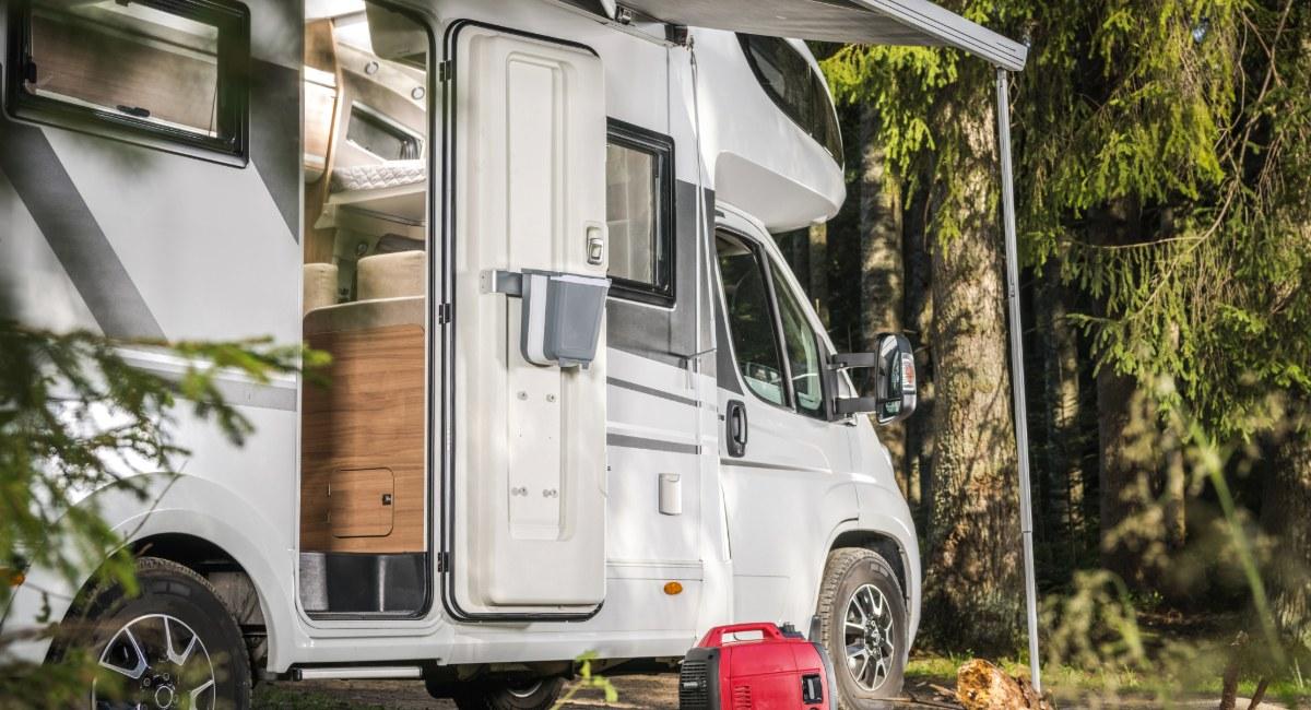 White Class C motorhome with open side door showing interior storage and wood cabinetry, red portable generator on ground, parked in wooded campsite setting ideal for family camping