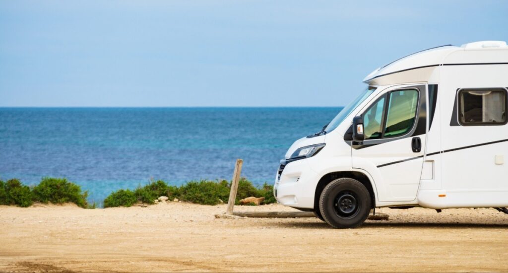White Class B camper van parked at scenic beachfront campsite with turquoise ocean water and coastal vegetation ideal for beach RV camping