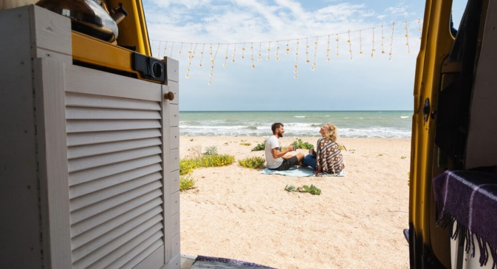 Young couple sitting on beach outside converted camper van enjoying oceanfront RV camping lifestyle and beach views
