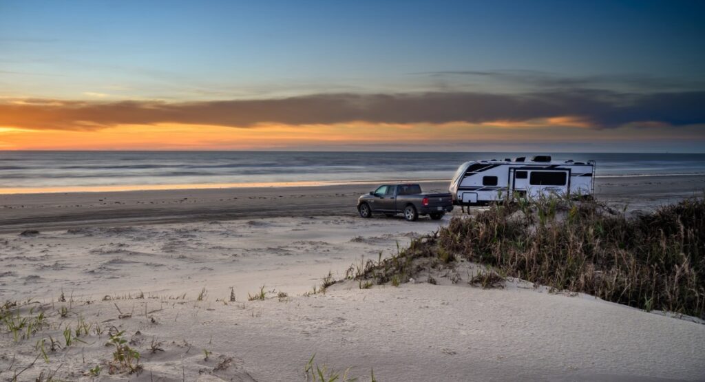 Truck towing travel trailer on sandy beach at sunset along Texas Gulf Coast with Pacific Ocean waves and dunes at Padre Island