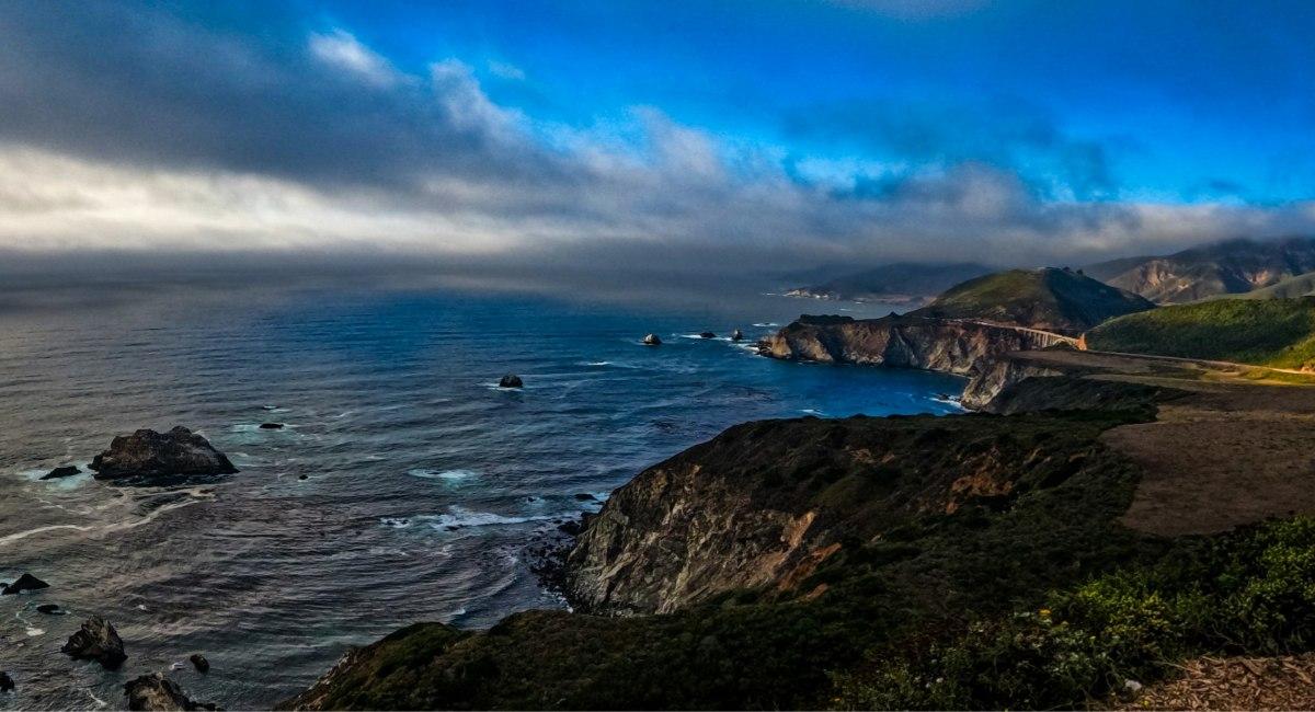 Dramatic panoramic view of the Big Sur coastline with rocky cliffs, sea stacks, and Bixby Bridge visible in the distance